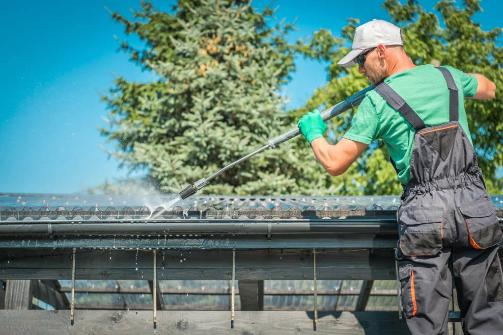A person cleans gutters on a sunny day, using a long pole to spray water