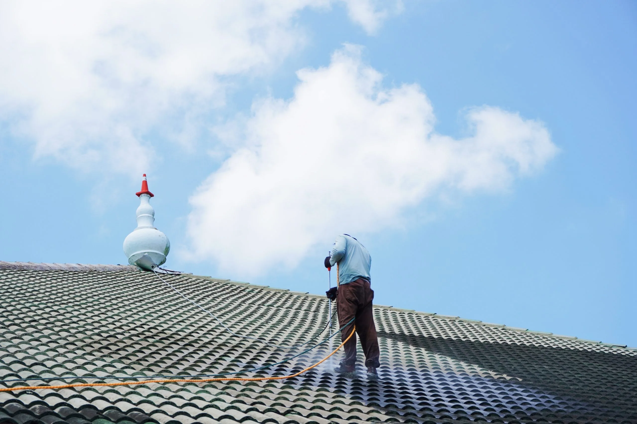 A person cleans a tiled roof under a clear blue sky with clouds