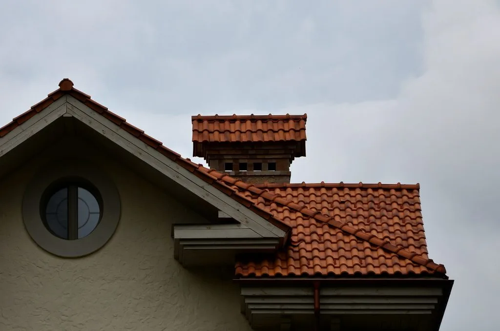 A sloped roof with red tiles and a decorative chimney against a cloudy sky